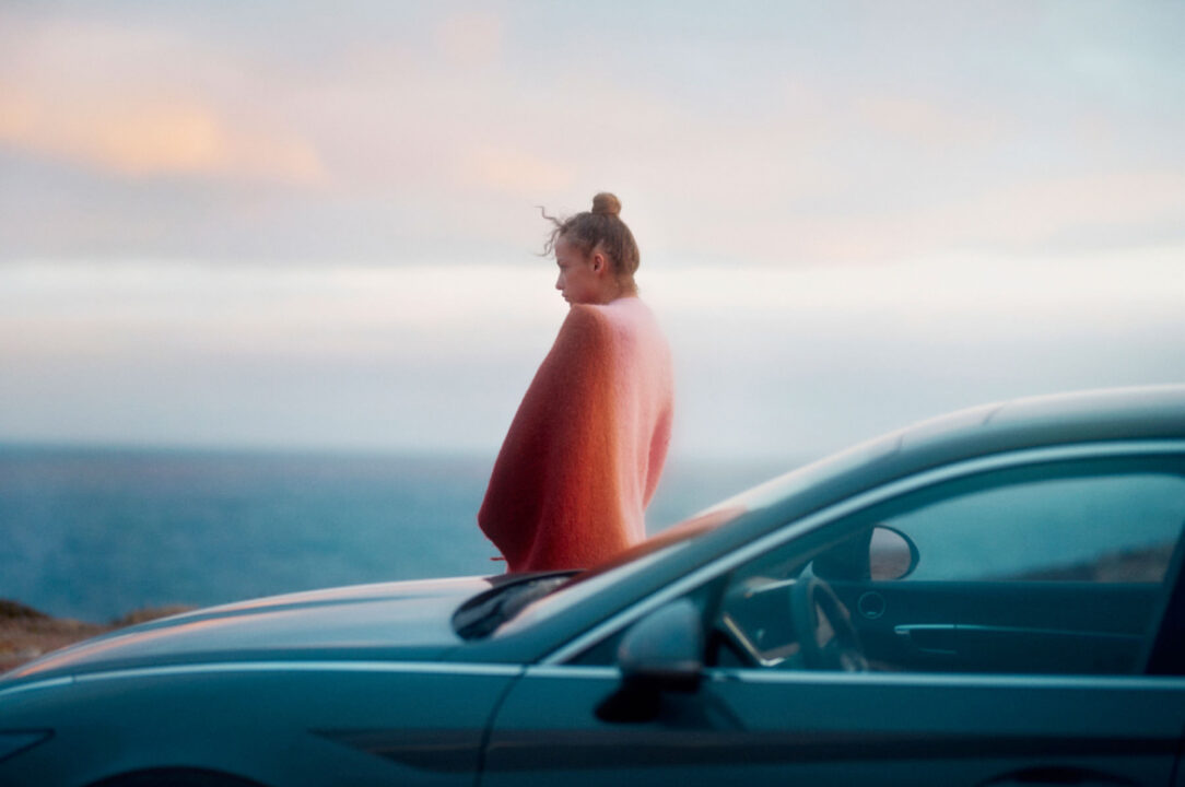 Eine Person, eingehüllt in eine rosa-rote Decke mit Farbverlauf, steht bei Sonnenuntergang am Meer neben einem blauen Auto, im Hintergrund ein sanft pastellfarbener Himmel.