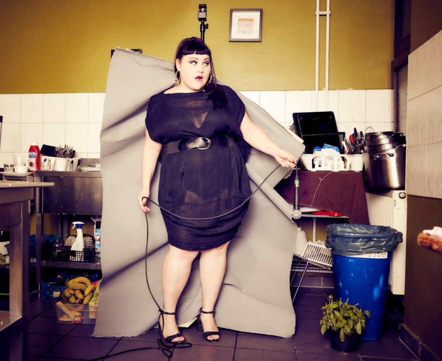 Eine Frau in einem schwarzen Kleid steht in einer Küche, die teilweise mit einem grauen Hintergrund verhüllt ist. Sie hält ein Kabel in der Hand. Im Hintergrund sind Besteck, Geschirr und ein Laptop zu sehen. Im Vordergrund sind ein blauer Mülleimer und eine Pflanze zu sehen.
