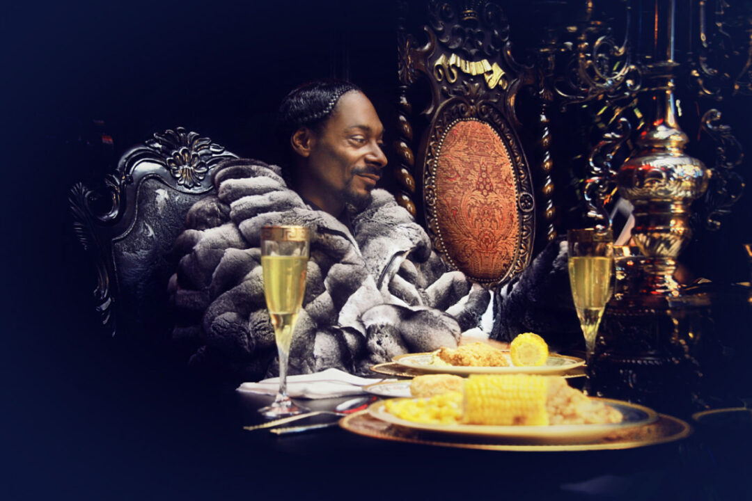 Ein Mann in einem luxuriösen Pelzmantel sitzt an einem reich verzierten Tisch mit goldenen Details und genießt in einem eleganten, schwach beleuchteten Ambiente eine Mahlzeit aus gebratenem Hühnchen, Maiskolben und Champagner.