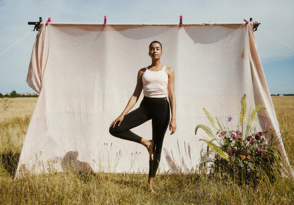 A person practices a yoga tree pose outdoors in front of a light pink fabric backdrop, with tall grass and flowers nearby under a clear sky.