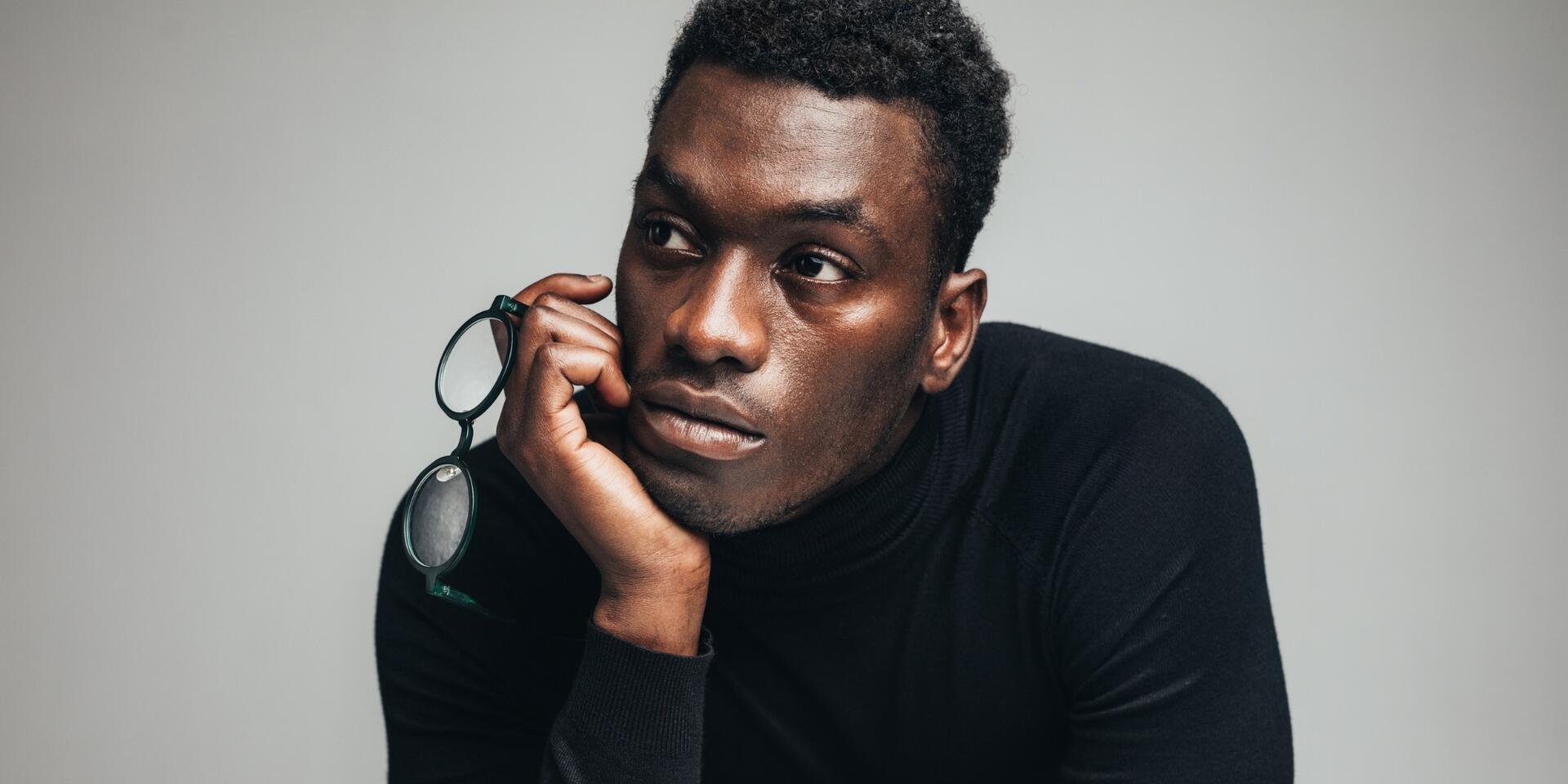 A man wearing a black turtleneck rests his chin on his hand while holding eyeglasses, looking thoughtfully into the distance against a plain gray background.