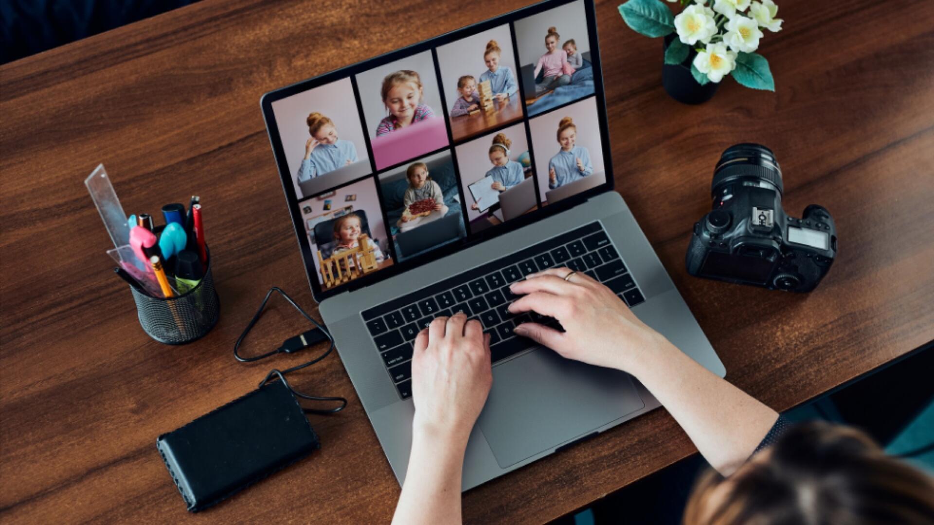 A person types on a laptop at a wooden desk during a video call with eight people. Nearby are a camera, pen holder, external drive, and a small potted plant with white flowers.