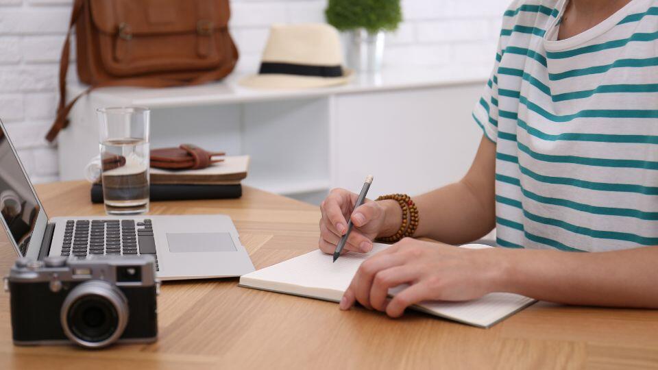 A person in a striped shirt writes in a notebook at a desk with a laptop, camera, glass of water, and wallet. Shelves in the background hold a hat, bag, and a potted plant.