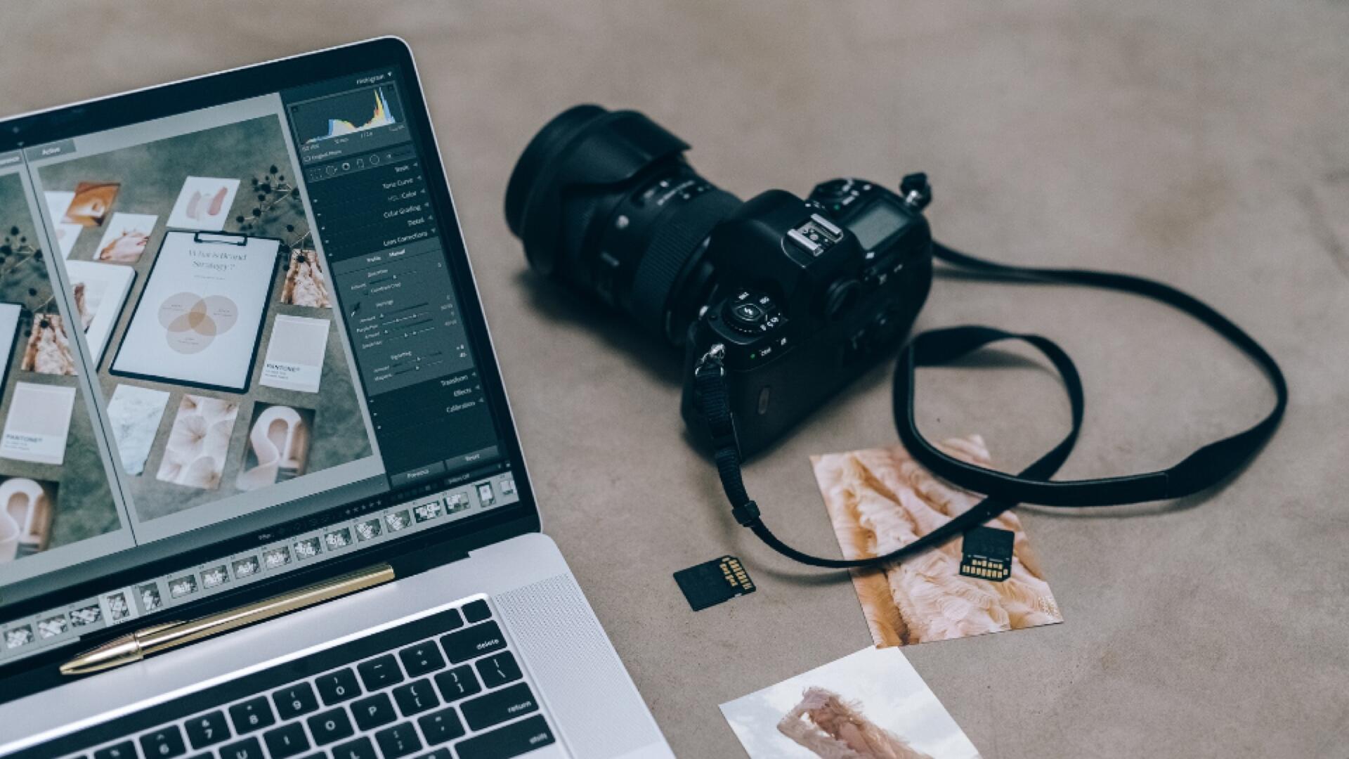 A laptop displaying photo editing software sits next to a DSLR camera, loose SD cards, and printed photographs on a beige surface.