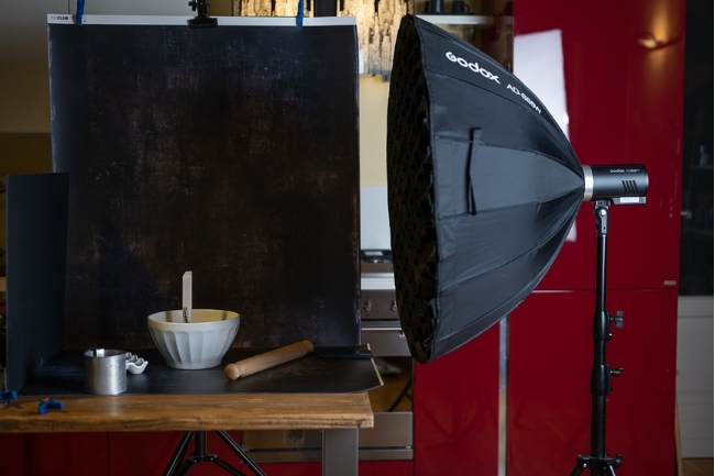 Behind-the-scenes food photography setup with a bowl and rolling pin on a table, a dark backdrop, and a large softbox light positioned beside the scene.