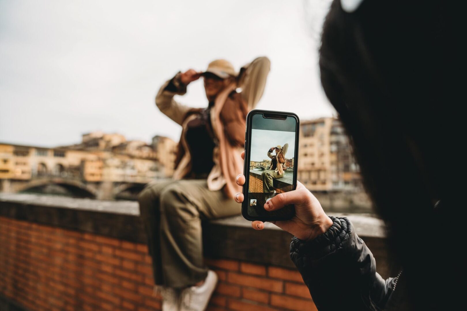 A person takes a photo with a smartphone of someone posing and adjusting their hat while sitting on a brick wall, with blurred buildings and a bridge in the background.