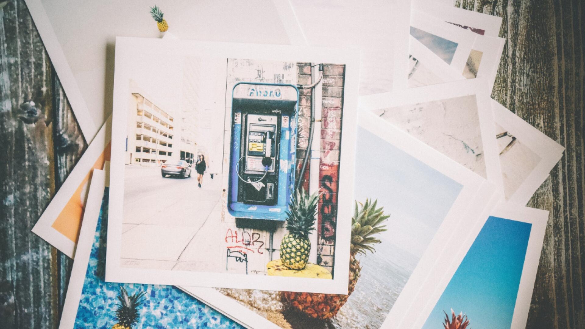 A stack of printed photographs lies on a wooden surface. The top photo shows a blue public payphone on a city street with graffiti. Other photos below feature pineapples, water, and beach scenes.