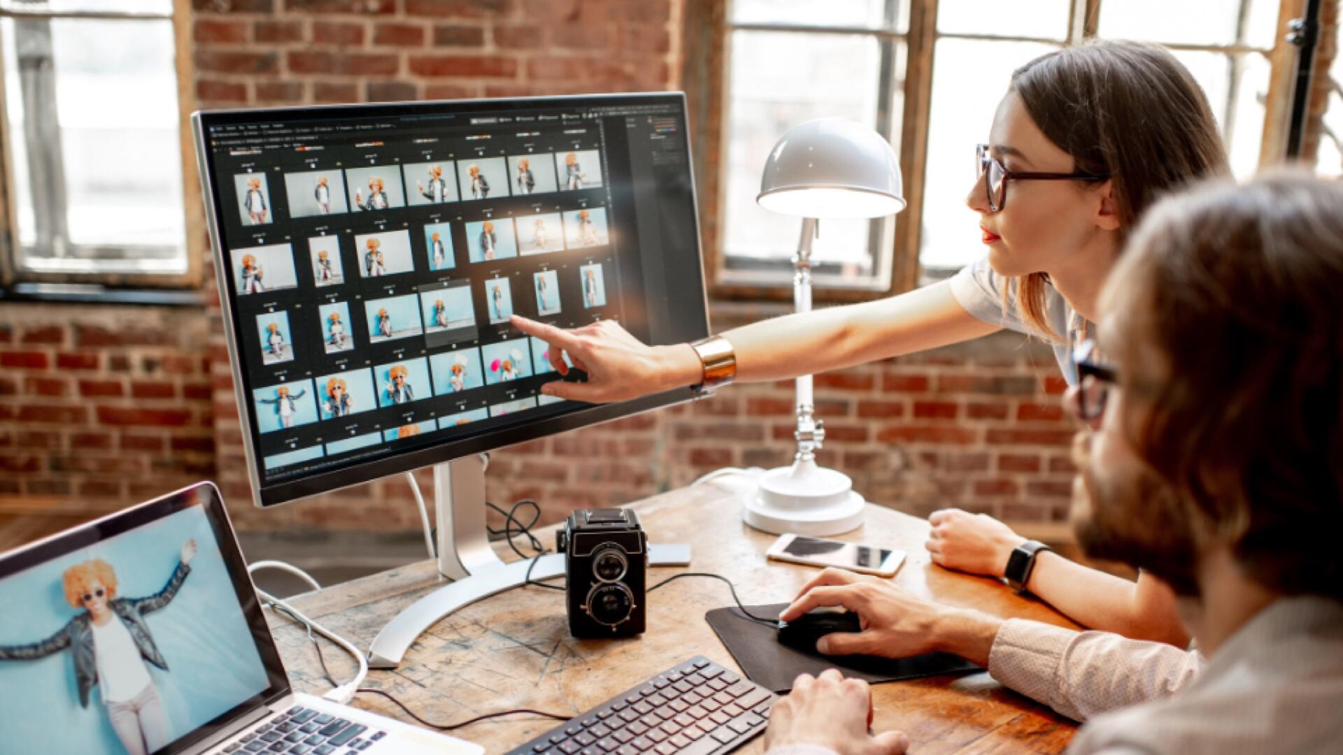 Two people work at a desk with a computer displaying thumbnails of photos. One person points at the screen while the other uses the mouse. A camera, lamp, and laptop are on the desk in a loft-style office.