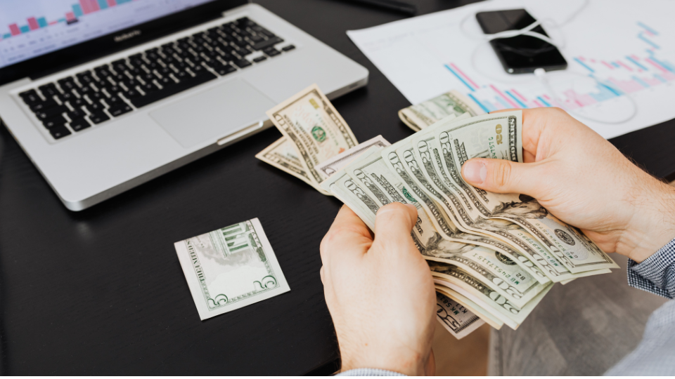 A person counts U.S. dollar bills at a desk with a laptop, smartphone, earphones, and financial charts.