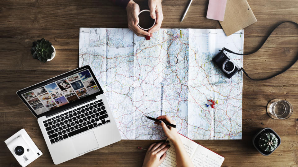 Two people plan a trip at a wooden table with a large map, laptop, camera, notebook, coffee cup, and a potted plant. One writes in a notebook while the other holds a cup over the map.