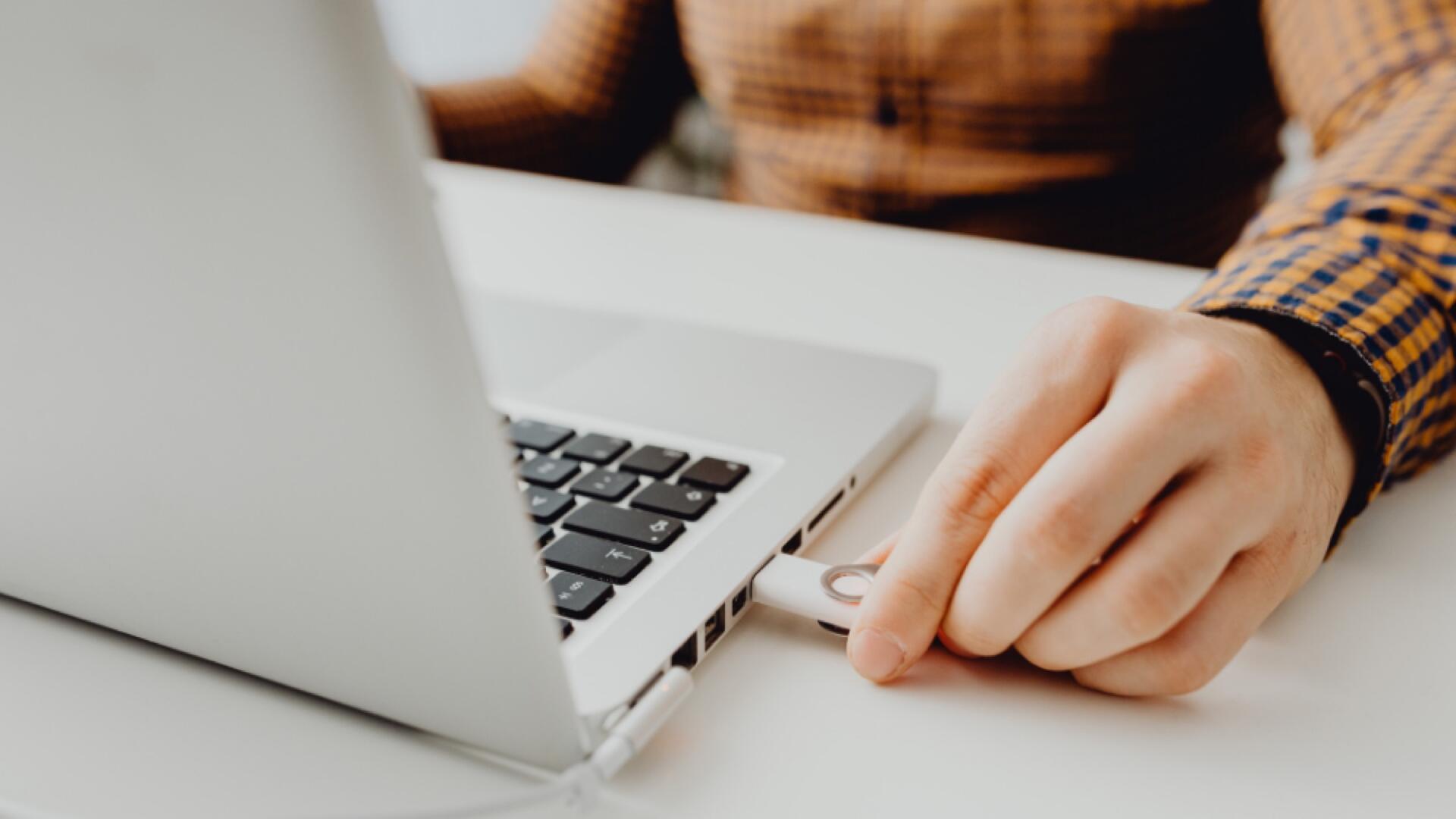 A person in a plaid shirt inserts a USB flash drive into a laptop on a white desk.