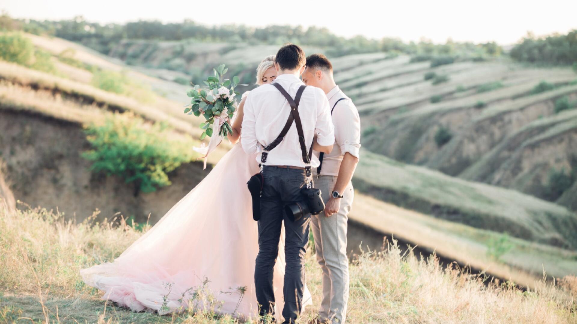 A bride and groom stand outdoors on a grassy hillside, holding hands and facing a photographer. The bride wears a long white dress and holds a bouquet; the men wear suspenders and light shirts. Scenic landscape in the background.