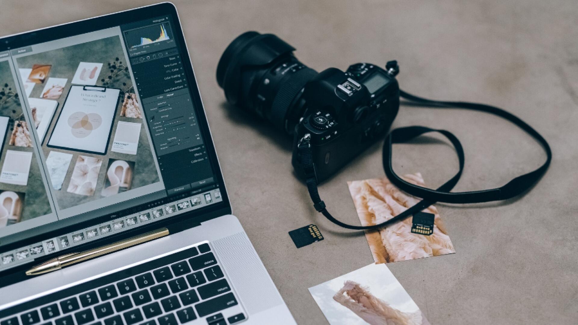 A laptop displaying photo editing software is next to a camera with a strap, two SD cards, a golden pen, and printed photos on a brown surface.