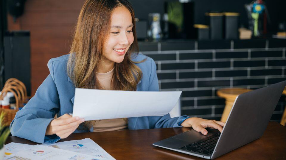 A woman sits at a desk, smiling and holding a sheet of paper while looking at her laptop. Several documents with charts and graphs are spread out on the desk in front of her.