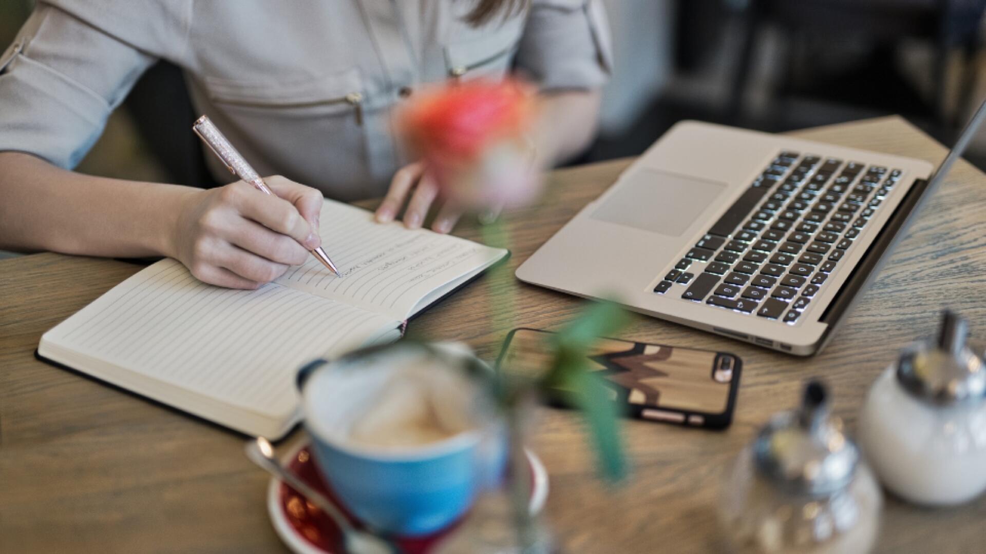 A person writes in a notebook at a wooden table with a laptop, a smartphone, a flower in a vase, a teacup, and a sugar bowl nearby. The setting appears to be a cozy café.